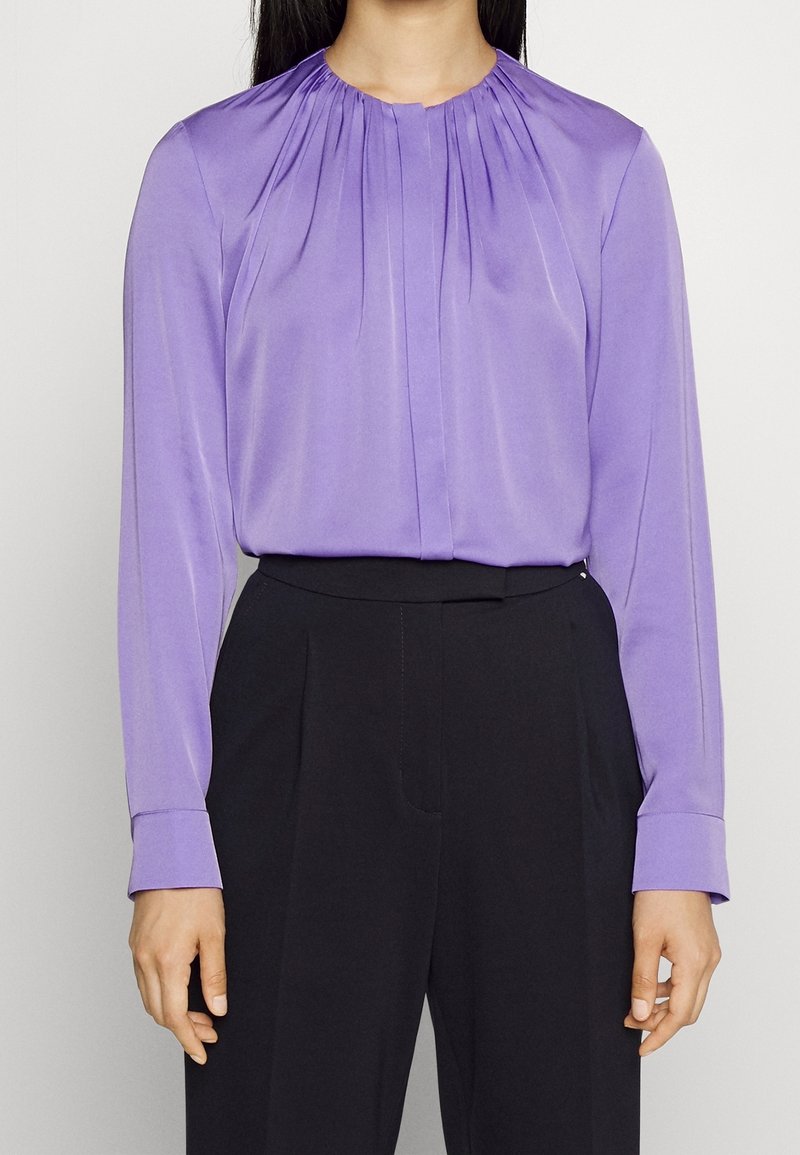 Femme portant un chemisier lavande plissé à manches longues, rentré dans un pantalon noir taille haute, vue des épaules aux cuisses.