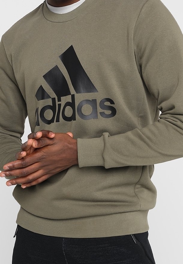 Homme portant un sweat-shirt Adidas vert olive avec un grand logo noir, mains jointes devant, associé à un pantalon noir.