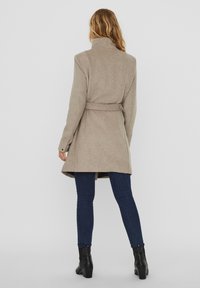 Beige ullblandad kappa med hög krage, bälte i midjan och långa ärmar. Har en texturerad yta och slutar under knäet. Återfinns tillsammans med mörka denim.