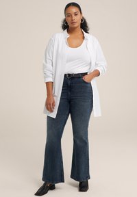 Camisa blanca de botones sobre una camiseta blanca sin mangas, combinada con jeans de campana azul oscuro y zapatos negros de tacón cuadrado. Incluye un cinturón negro simple.