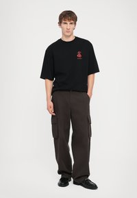 Schwarzes T-Shirt mit kurzen Ärmeln und roter Grafik, kombiniert mit locker sitzenden, dunkelbraunen Cargo-Hosen, die über Seitentaschen und einen strukturierten Stoff verfügen.