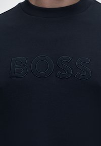 Gros plan d'un sweat-shirt bleu marine à col rond avec le mot « BOSS » brodé en fil assorti sur la poitrine.