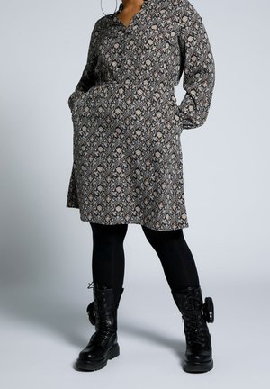 Femme portant une robe noire à motifs, des leggings noirs et des bottines noires à lacets avec poches latérales, debout les mains dans les poches de la robe.