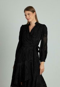Schwarzes Wickelkleid aus strukturiertem Stoff mit floralen Mustern, langen Ärmeln und einem Bindegürtel in der Taille. Der asymmetrische Saum verleiht Bewegung.