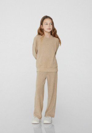 Beige sweatshirt and matching wide-leg pants made of soft, textured fabric. Model stands with arms behind, wearing white sneakers.