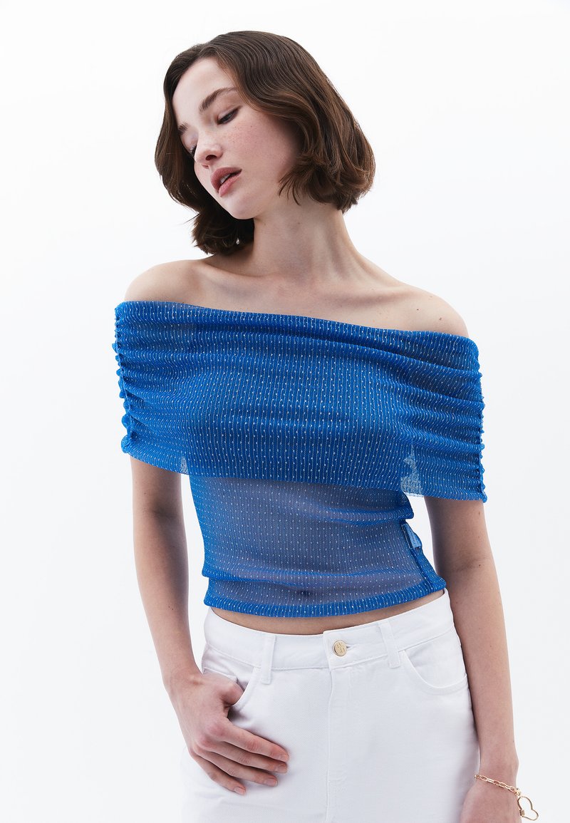 Modrá top s odkrytými rameny a řasenou strukturou, s metalickými akcenty a průsvitným designem, v kombinaci s bílými kalhotami vysokého pasu.