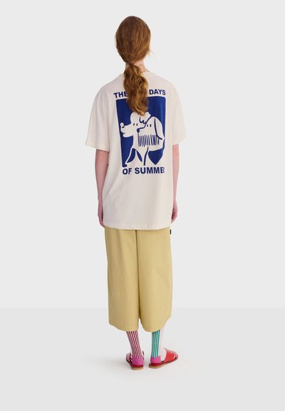 Personne aux cheveux longs attachés portant un t-shirt crème avec un motif de chien bleu et du texte, un pantalon large beige, des chaussettes rayées dépareillées et des sandales rouges.