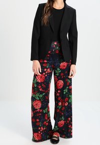 Zwarte op maat gemaakte blazer over een zwart topje, gecombineerd met wijde broek met een multikleurig bloemenpatroon op een donkere achtergrond. Zwarte schoenen.