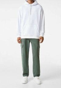 Vit hoodie med ficka fram, i kombination med gröna cargobyxor med sidfickor och vita texturerade sneakers.