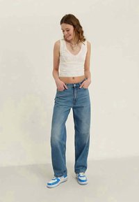 Giovane donna che indossa una maglietta bianca senza maniche, jeans blu larghi e sneaker blu e bianche, in piedi con le mani nelle tasche, guardando in basso e sorridendo.