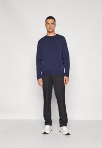 Ναυτικό φανελάκι με μακριά μανίκια και λαιμόκοψη, που διαθέτει ένα μικρό λογότυπο σε μπλε χρώμα. Συμπληρώνεται με σκούρες παντελόνες και λευκά αθλητικά παπούτσια.