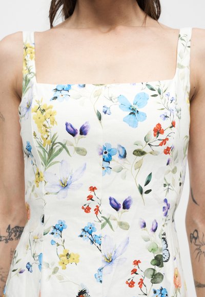 Γυναίκα που φοράει λευκό φόρεμα με τετράγωνο ντεκολτέ, διακοσμημένο με πολύχρωμα λουλουδένια και πεταλούδες σχέδια, δείχνοντας τατουάζ και στα δύο χέρια.