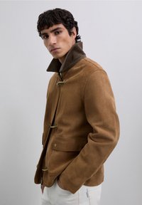 Chaqueta de ante marrón con un cuello de pana, que presenta cierres de tipo toggle y bolsillos laterales, silueta estructurada y textura suave.