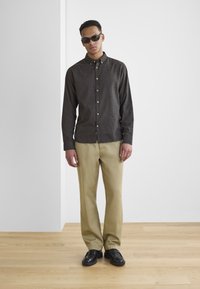 Brown button-up shirt with a subtle texture, paired with beige pants and black shoes. The model is wearing dark sunglasses.