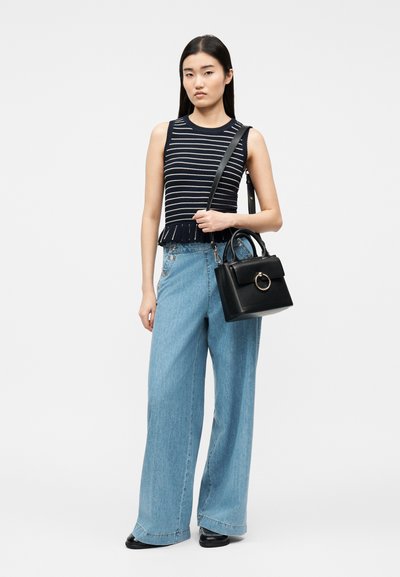 Γυναίκα με μπλε ριγέ τοπ χωρίς μανίκια και τζιν παντελόνι με φαρδύ πόδι, κρατώντας μαύρη τσάντα με κυκλική μεταλλική λεπτομέρεια, στέκεται μπροστά σε ανοιχτό φωτεινό φόντο.