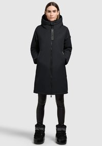 Zwarte geïsoleerde jas met een grote capuchon, voorrits en zijzakken. Draagt over zwarte leggings en kniehoge zwarte laarzen.