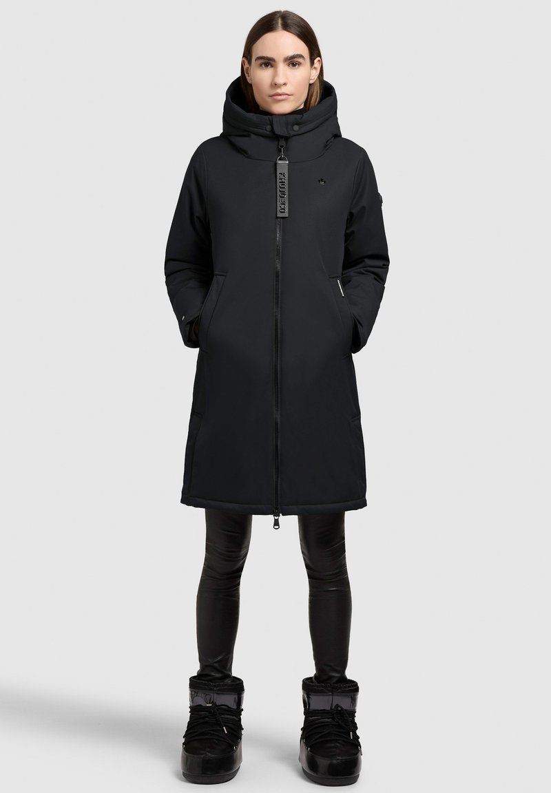 Zwarte geïsoleerde jas met een grote capuchon, voorrits en zijzakken. Draagt over zwarte leggings en kniehoge zwarte laarzen.