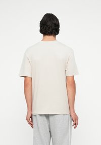 Lys beige bomulds t-shirt med en afslappet pasform, korte ærmer og rund halsudskæring, vist fra ryggen mod en ensfarvet baggrund.