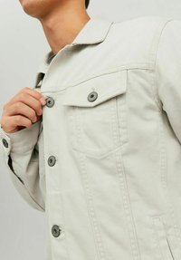 Giacca in denim grigio chiaro con colletto classico, due tasche sul petto e bottoni a pressione neri. Il tessuto ha una finitura testurizzata.