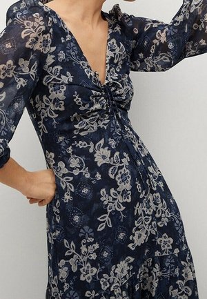 Kobieta ubrana w granatową sukienkę w kwiatowy wzór z marszczonym przodem i przezroczystymi rękawami o długości do łokcia, pozująca z jedną ręką na biodrze.