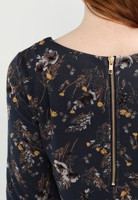 Blommönstrad mörkblå klänning med rund halsringning och en gulddragkedja bak, med inslag av gula och vita blommor.