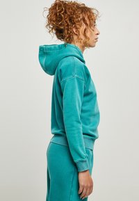 Tirkīza krāsas kapuccīna krekls, izgatavots no kokvilnas maisījuma, ar aukliņu kapuci, ribotām piedurknēm un brīvu piegriezumu, kopā ar atbilstošām sporta biksēm.