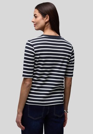 Žena s dlouhými tmavými vlasy, oblečená v černobílém proužkovaném tričku s krátkými rukávy a tmavě modrých džínách, stojící před jednobarevným šedým pozadím.