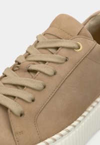 Světlý semišový tenisk s kulatou špičkou, béžovými tkaničkami, zlatými očky a bílou gumovou podrážkou s texturovaným okrajem. Hladký povrch a minimalistický design.