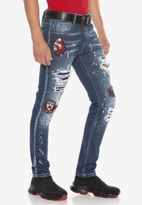 Jean skinny en denim bleu foncé, avec des déchirures usées, des éclaboussures de peinture blanche et divers patchs brodés. Associé à des baskets noires à clous.