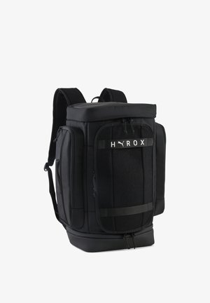 Czarny plecak Puma z wieloma zapinanymi na zamek komorami, paskami na ramiona oraz logo "HYROX" na przedniej kieszeni.