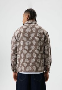 Veste marron à motif en tissu texturé, avec un col, des manches longues et un design ouvert orné de motifs tourbillonnants blancs.