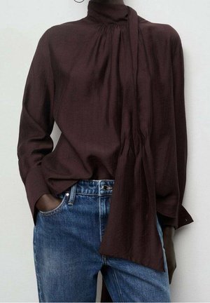 Dark brown long-sleeve blouse with high neck and extended scarf detail paired with blue jeans, one hand tucked in pocket, neutral background.