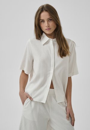 Žena s dlhými hnedými vlasmi má na sebe voľnú bielu krátku košeľu na gombíky a zodpovedajúce biele nohavice s elastickým pásom, stojí s jednou rukou v vrecku.