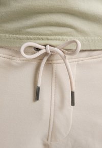 Gros plan sur un pantalon de survêtement beige clair avec un cordon de serrage noué au-dessus d'une taille côtelée et un ourlet supérieur vert olive doux visible.