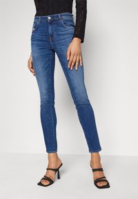 Úzké modré džíny vyrobené z denimu, s mírným pasem, designem s pěti kapsami a jemným vyblednutím. Černé sandály na vysokém podpatku doplňují vzhled.