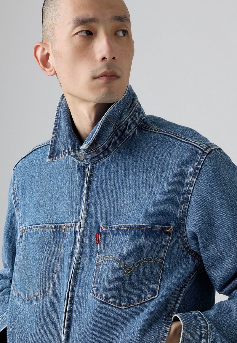 Giacca di denim blu con colletto alto, due tasche sul petto, cuciture a contrasto e dettaglio con etichetta del brand. Indossata da una persona con capelli corti.