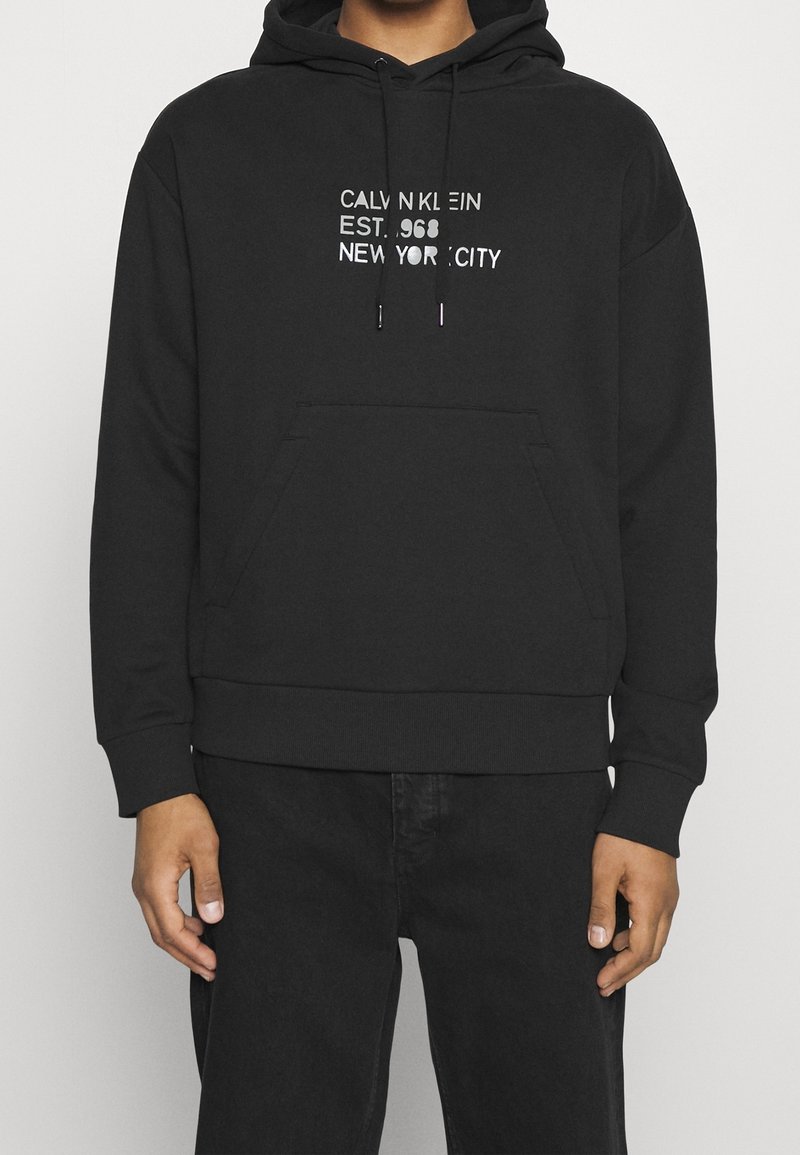Sweat à capuche noir avec poche avant et texte blanc indiquant "CALVIN KLEIN EST. 1968 NEW YORK CITY,", porté par une personne les mains relâchées le long du corps.