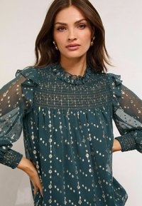 Zöldes-kék blúz fodros díszítéssel, smockolt mellrésszel és áttetsző ujjakkal. Fémes csillogással díszítve, hogy fokozza a textúrát.