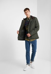 Quilted green jacket with brown collar, front pockets, and golden snaps, layered over a plaid shirt, paired with blue jeans and white shoes.