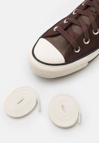 Braune Wildleder-Sneaker mit weißer Gummikappe und schwarzem Sohlenrand, ausgestattet mit braunen Schnürsenkeln. Zwei weiße runde Schnürsenkel mit Plastikkappen liegen daneben.