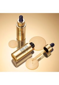 To guldflasker med Estee Lauder serum og pipetter, den ene delvist åben, omgivet af geldråber på en reflekterende guldoverflade.