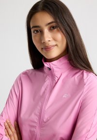Ljuskorrig pink vindjacka med hög krage och dragkedja framtill. Slät textur och logodetalj på vänster bröst.