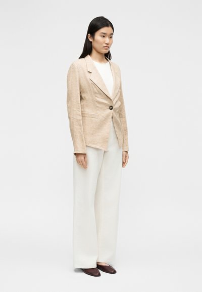 Γυναίκα όρθια φορώντας μπεζ σακάκι με ένα κουμπί, λευκό παντελόνι με φαρδιές μπατζάκια και καφέ μυτερές παπούτσια σε απλό φωτεινό φόντο.