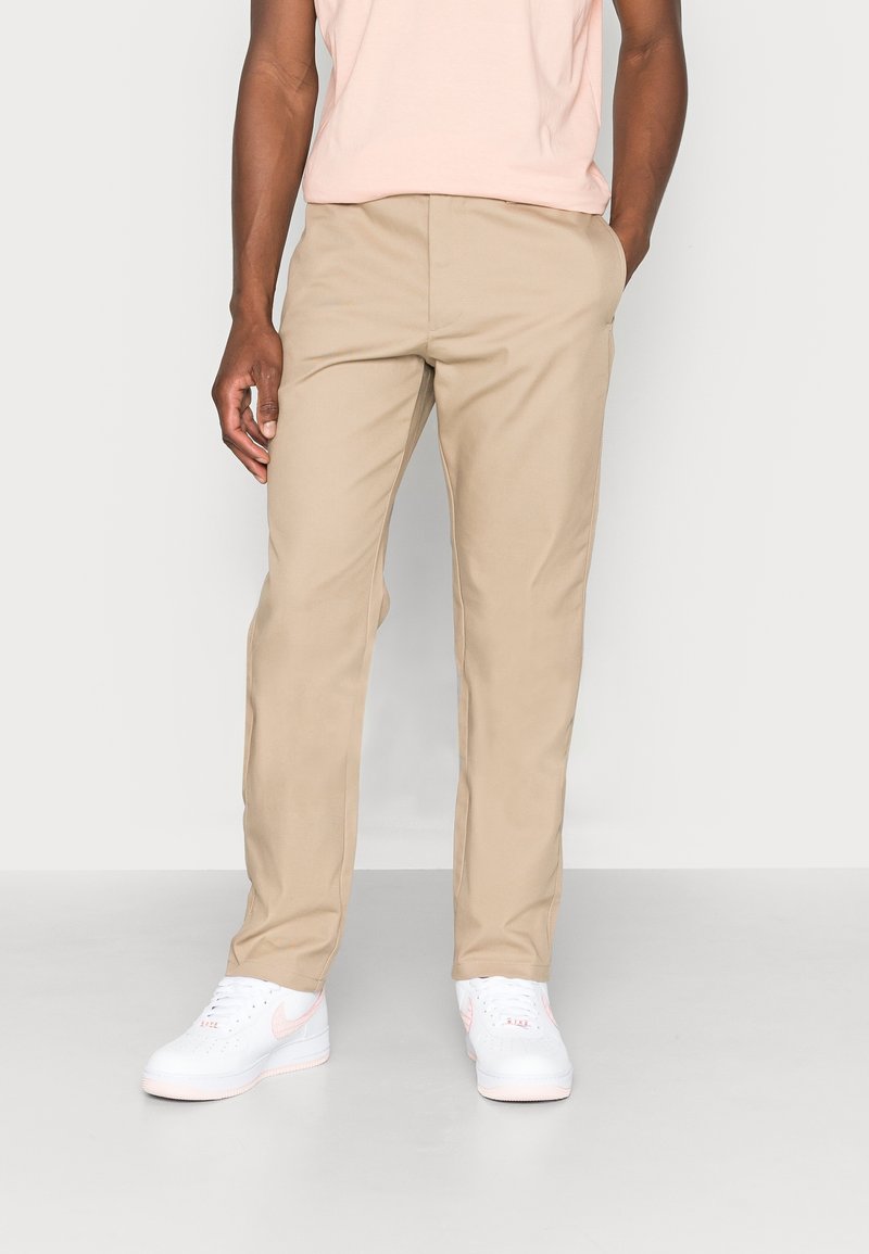 Chinos beiges en tissu mélange de coton, coupe droite, avec poches latérales, associés à des baskets blanches avec des accents roses.