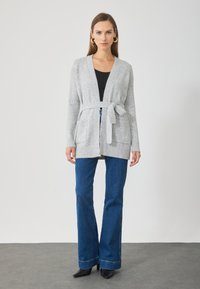 Pilka mezgimo kardiganas su raiščiu juosmenyje, V formos iškirpte ir dviem priekinėmis kišenėmis, derinama su tamsiai mėlynomis plačiomis džinsomis ir juodais aukštakulniais bateliais.