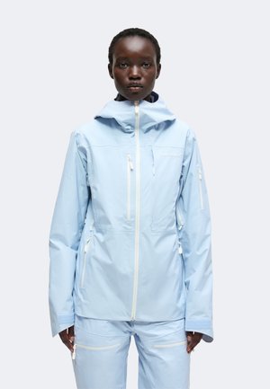 Person trägt eine hellblaue wasserdichte Jacke und passende Hose mit mehreren weißen Reißverschlüssen vor einem schlichten hellen Hintergrund.