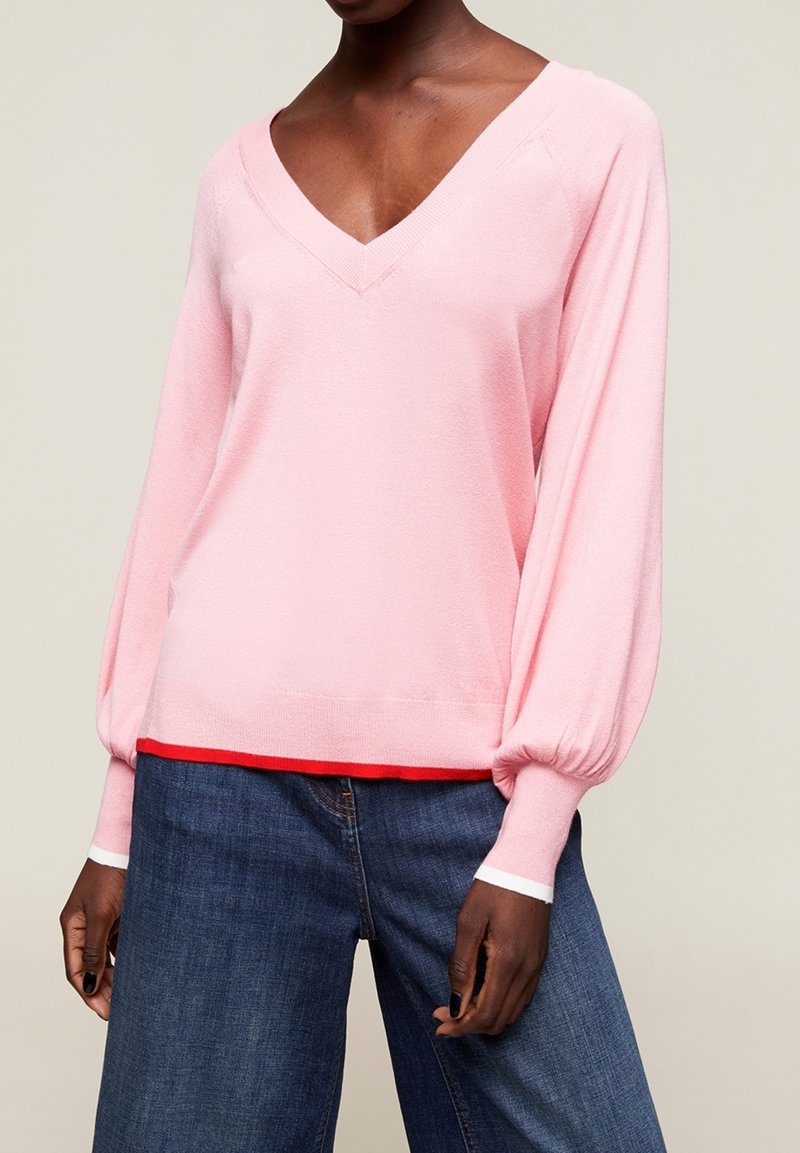 Rosa stickad tröja med djup V-hals, långa ballongenärmar och ribbad nederkant. Lagd över en röd skjorta, tillsammans med mörk denimjeans.