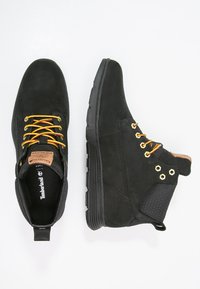 Botines de ante negros con cordones amarillos y rojos, ojales dorados, acentos texturizados y suela de goma. Presenta una etiqueta de marca de color marrón claro en la lengüeta.