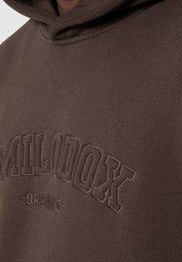 Sudadera marrón con una textura suave, que presenta un diseño de logo en relieve que dice "MILODOX ORIGINALS" en la parte frontal.