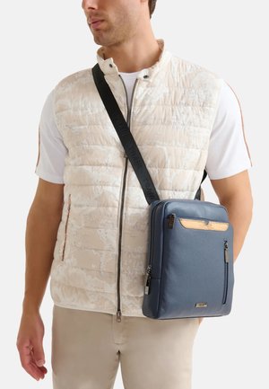 Borsa a tracolla in pelle blu con tasca frontale con zip, finitura testurizzata e accentuazione marrone chiaro nella parte superiore. Indossata con un gilet chiaro e una camicia bianca.